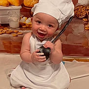 Rory joined the competition — help win amazing prizes! baby, chef_hat, apron, smiling, kitchen, bread, pastries, eggs, jar, bagels, cooking_utensil, sitting, white_cloth, cute, child, person, indoors, food, playful, portrait
