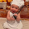 baby, chef_hat, apron, smiling, kitchen, bread, pastries, eggs, jar, bagels, cooking_utensil, sitting, white_cloth, cute, child, person, indoors, food, playful, portrait
