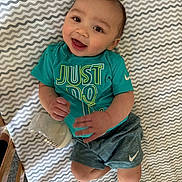 Rory is registered to the contest to win money with this photo: baby, infant, smiling, bottle, clothing, teal_shirt, shorts, sock, lying_down, patterned_blanket, happy, cute, person, indoor, wooden_crib, milk_bottle, face, skin, head, hand