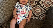 Jocelyn is registered to the contest to win money with this photo: baby, child, tongue_out, onesie, socks, couch, cushion, patterned_cushion, indoor, smiling, seated, cheeky, cute, infant, person, furniture, home, relaxed, happy, portrait