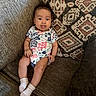 baby, child, tongue_out, onesie, socks, couch, cushion, patterned_cushion, indoor, smiling, seated, cheeky, cute, infant, person, furniture, home, relaxed, happy, portrait