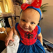 Nina is registered to the contest to win money with this photo: baby, blue_dress, child, cute, eyes, face, furniture, hand, happy, headband, holding, home, indoor, person, portrait, red_bow, smiling, spiderman_dress, toddler, wall_art