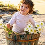child, toddler, curly_hair, bucket, flowers, daisies, outdoor, sunlight, nature, smile, white_clothing, rustic, wood, greenery, cute, baby, portrait, playful, garden, happy