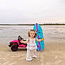 toddler, child, beach, sand, toy_car, surfboard, curly_hair, blue_surfboard, pink_toy_car, yellow_toy, outdoor, water, sky, cloudy, shoreline, standing, clothing, cute, small_person, playtime