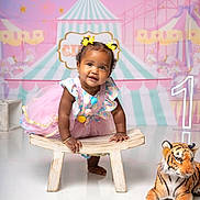 Willow joined the competition — help win amazing prizes! baby, toddler, girl, smiling, crawling, colorful_clothing, tutu, clown_outfit, yellow_bows, wooden_bench, plush_tiger, birthday, number_one, carnival_background, carousel, stars, pastel_colors, indoor, cute, celebration