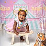 baby, toddler, girl, smiling, crawling, colorful_clothing, tutu, clown_outfit, yellow_bows, wooden_bench, plush_tiger, birthday, number_one, carnival_background, carousel, stars, pastel_colors, indoor, cute, celebration