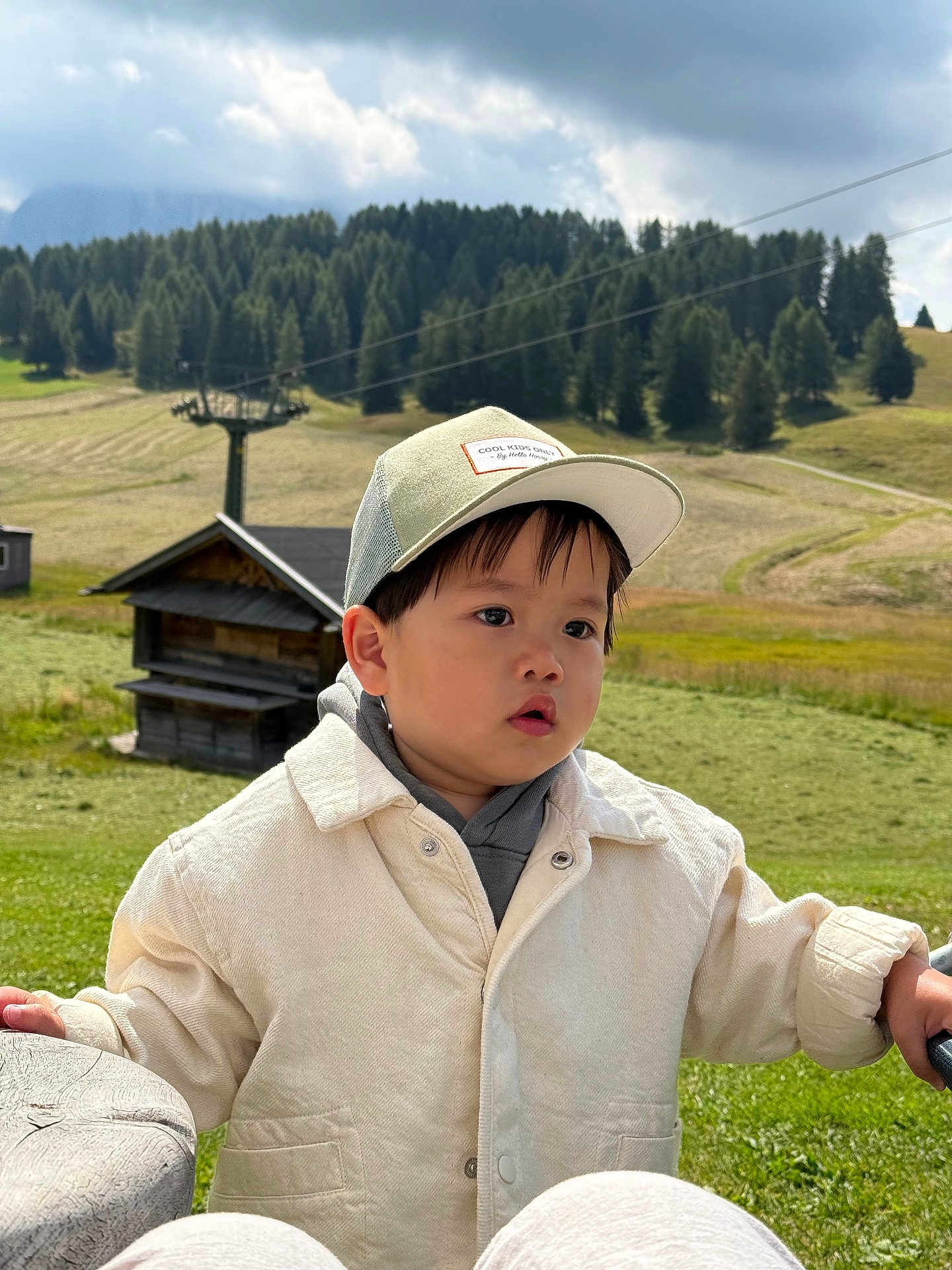 Kei a rejoint le concours — aidez-le/la à gagner de superbes lots ! child, toddler, boy, cap, jacket, outdoor, mountains, grass, trees, wooden_structure, sky, clouds, nature, scenic, daylight, portrait, curious, casual_clothing, greenery, person