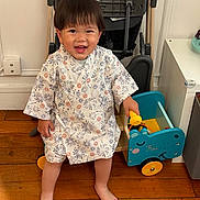 Kei a rejoint le concours — aidez-le/la à gagner de superbes lots ! toddler, child, smiling, barefoot, toy, robe, wooden_floor, indoor, stroller, blue_toy_cart, happy, person, young_child, clothing, furniture, playtime, home, floor, cute, standing