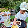 child, toddler, cap, food, chips, cake, drink, lemon, glass, outdoor, greenery, grass, table, tray, adult, relaxing, nature, snack, summer, daytime