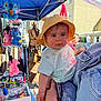 baby, child, yellow_hat, white_shirt, denim_jacket, outdoor, market, stall, plush_toys, accessories, hand, watch, people, shopping, bags, colorful, blue_canopy, curious, person, portrait
