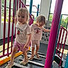 alphabet_board, blonde_hair, blue_poles, casual_clothing, children, climbing_structure, daylight, indoor_playground, pink_railings, play_equipment, playground, shorts, siblings, smiling, sneakers, standing, t_shirts, toddlers, walking, windows