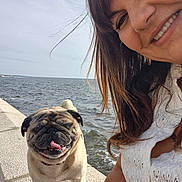 Pablo participe au concours pour gagner de l'argent avec cette photo : dog, pug, smiling, woman, selfie, sea, water, outdoor, daylight, tongue_out, stone_walkway, happy, pet, casual_clothing, short_hair, accessories, nature, portrait, sunlight, leisure