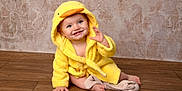Freya is registered to the contest to win money with this photo: baby, child, yellow_robe, duck_hood, smiling, waving, barefoot, soft_toy, wooden_floor, indoor, portrait, cute, happy, toddler, plush_toy, warm_clothing, cozy, infant, sitting, background_wallpaper