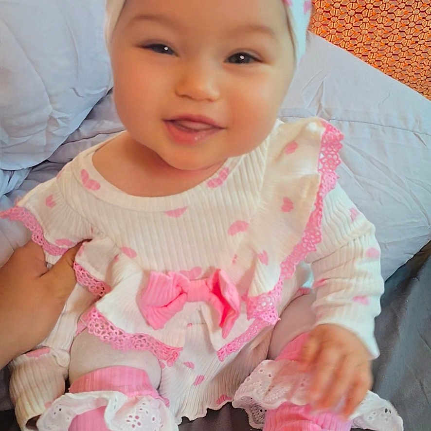 Nayra participe au concours pour gagner de l'argent avec cette photo : baby, child, smiling, pink, white, headband, bed, pillow, patterned_wall, clothing, happy, cute, infant, sitting, indoors, portrait, soft_lighting, hand, fabric, cozy