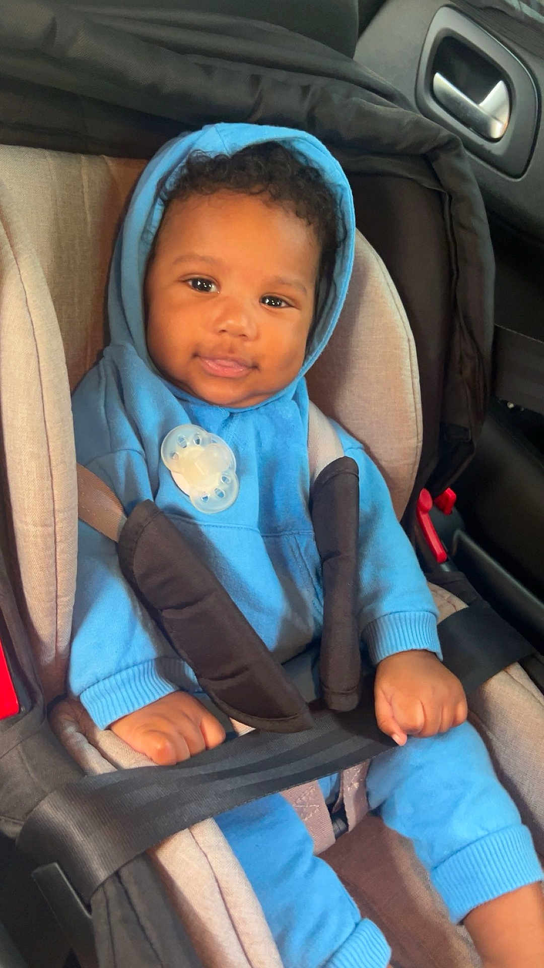 Lorenzo participe au concours pour gagner de l'argent avec cette photo : baby, child, car_seat, blue_hoodie, pacifier, seatbelt, smiling, curly_hair, infant, cute, indoors, car_interior, safety, young_child, clothing, hands, face, portrait, seated, casual
