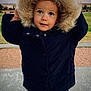 child, coat, hood, fur, winter_clothing, outdoor, park, grass, sidewalk, curious, face, person, cold_weather, jacket, blue_pants, young, cute, portrait, daylight, nature