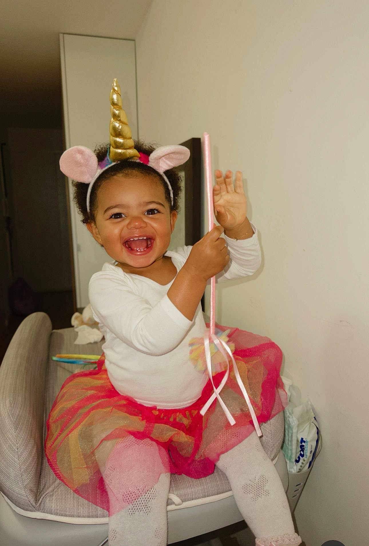 Beatrice participe au concours pour gagner de l'argent avec cette photo : child, toddler, smile, unicorn_headband, wand, tutu, sitting, indoor, happy, curly_hair, white_top, pink_tights, chair, playful, toy, portrait, accessory, room, baby_wipes, laughing