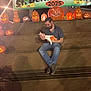 baby, man, pumpkin, jack_o_lantern, sign, wooden_bleachers, night, festival, holding, orange_clothing, white_clothing, smiling, halloween, light, shadow, outdoor, event, celebration, person, bench