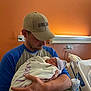man, baby, newborn, hospital_room, blanket, cap, holding, father, parent, bed, cup, table, clothing, person, indoor, medical_equipment, facial_hair, hat, sleeping, skin