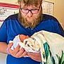 man, baby, newborn, glasses, beard, blue_shirt, blanket, indoor, holding, portrait, tender, hospital, health_poster, person, face, male, newborn_hat, hands, wall, closeup
