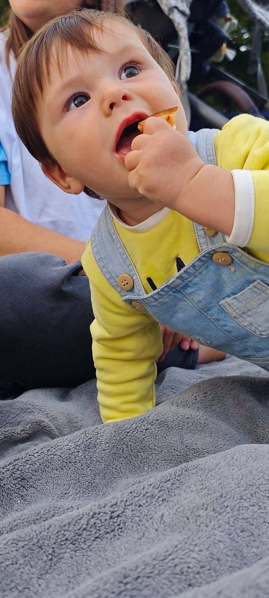 baby, blanket, candid, casual_clothing, child, curious, daylight, denim_overalls, eating, face, grass_background, hand, outdoor, person, portrait, relaxed, snack, toddler, wide_eyes, yellow_shirt
