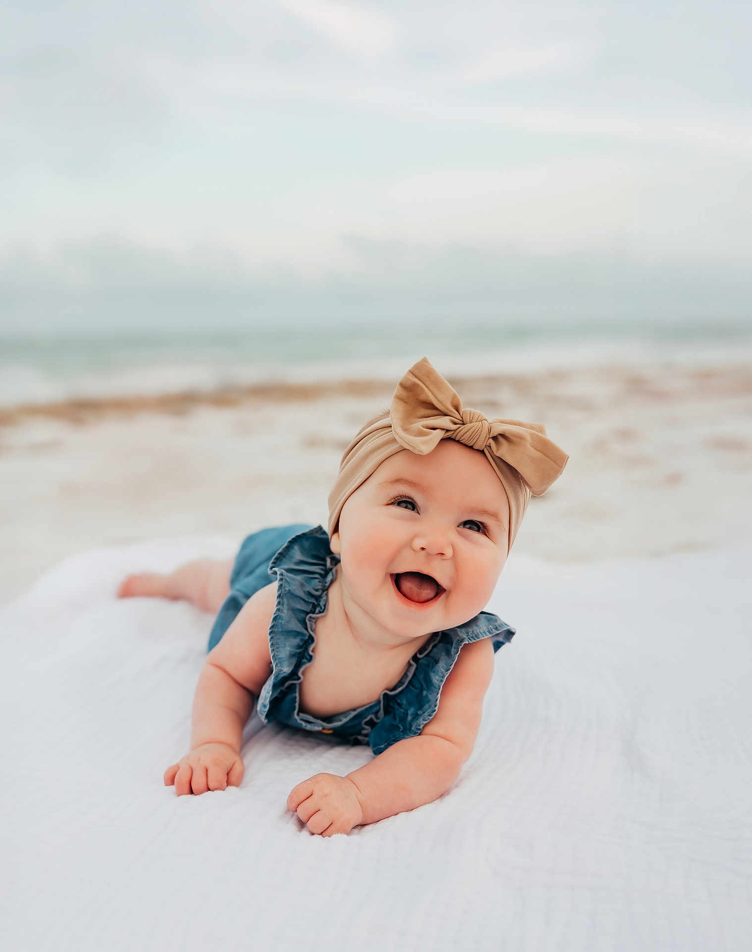 Emma joined the competition — help win amazing prizes! baby, infant, smiling, headband, bow, denim, outdoor, beach, blanket, lying_down, happy, child, cute, portrait, face, skin, natural_light, ocean, sky, blurred_background