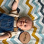 baby, infant, child, denim_overalls, beige_shirt, blue_eyes, smile, arm_raised, zigzag_pattern, colorful_blanket, wooden_plaque, milestone, one_month, lying_down, cute, portrait, indoors, soft_lighting, happy, person