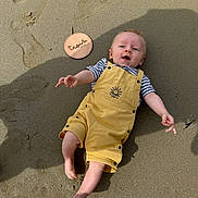 Malo a rejoint le concours — aidez-le/la à gagner de superbes lots ! baby, child, sand, yellow_overalls, striped_shirt, barefoot, smiling, shadow, wooden_plaque, age_marker, outdoor, sunny, happy, cute, infant, reaching, skin, footprint, playful, summer