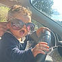 child, toddler, sunglasses, car, steering_wheel, curly_hair, hand, sunlight, window, seat, clothing, person, portrait, expression, vehicle_interior, daylight, outdoor, reflection, casual, cute