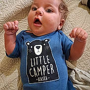 William joined the competition — help win amazing prizes! baby, child, onesie, blue_clothing, curly_hair, face, expression, quilt, bed, raised_fists, indoor, portrait, cute, infant, person, text_on_clothing, clothing, happy, young_child, lying_down