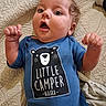 baby, child, onesie, blue_clothing, curly_hair, face, expression, quilt, bed, raised_fists, indoor, portrait, cute, infant, person, text_on_clothing, clothing, happy, young_child, lying_down