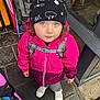 backpack_straps, beanie, child, cold_weather, curious_expression, curly_hair, green_eyes, looking_up, outdoor, pavement, person, pink_jacket, raincoat, rainy_day, standing, street, umbrella, urban, wet_pavement, white_shoes