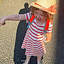 child, curly_hair, fashion, flower, greenery, happy, holding_hand, nature, outdoor, paved_path, person, playful, road, shadow, silver_sandals, smiling, straw_hat, striped_dress, summer, sunlight