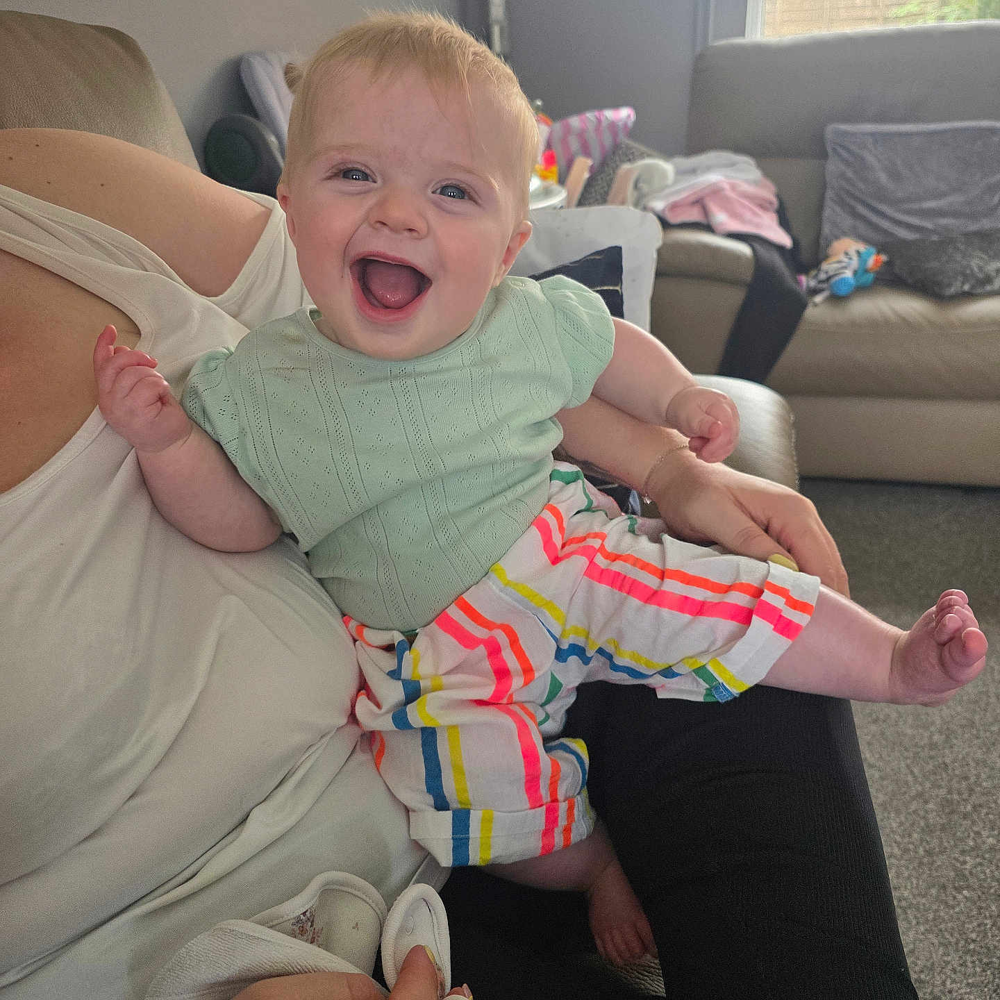 Scarlett joined the competition — help win amazing prizes! adult_arm, baby, carpet, casual, child, colorful_clothing, couch, cute, furniture, happy, indoor, joy, living_room, mint_green_top, natural_light, person, portrait, smiling, striped_pants, window