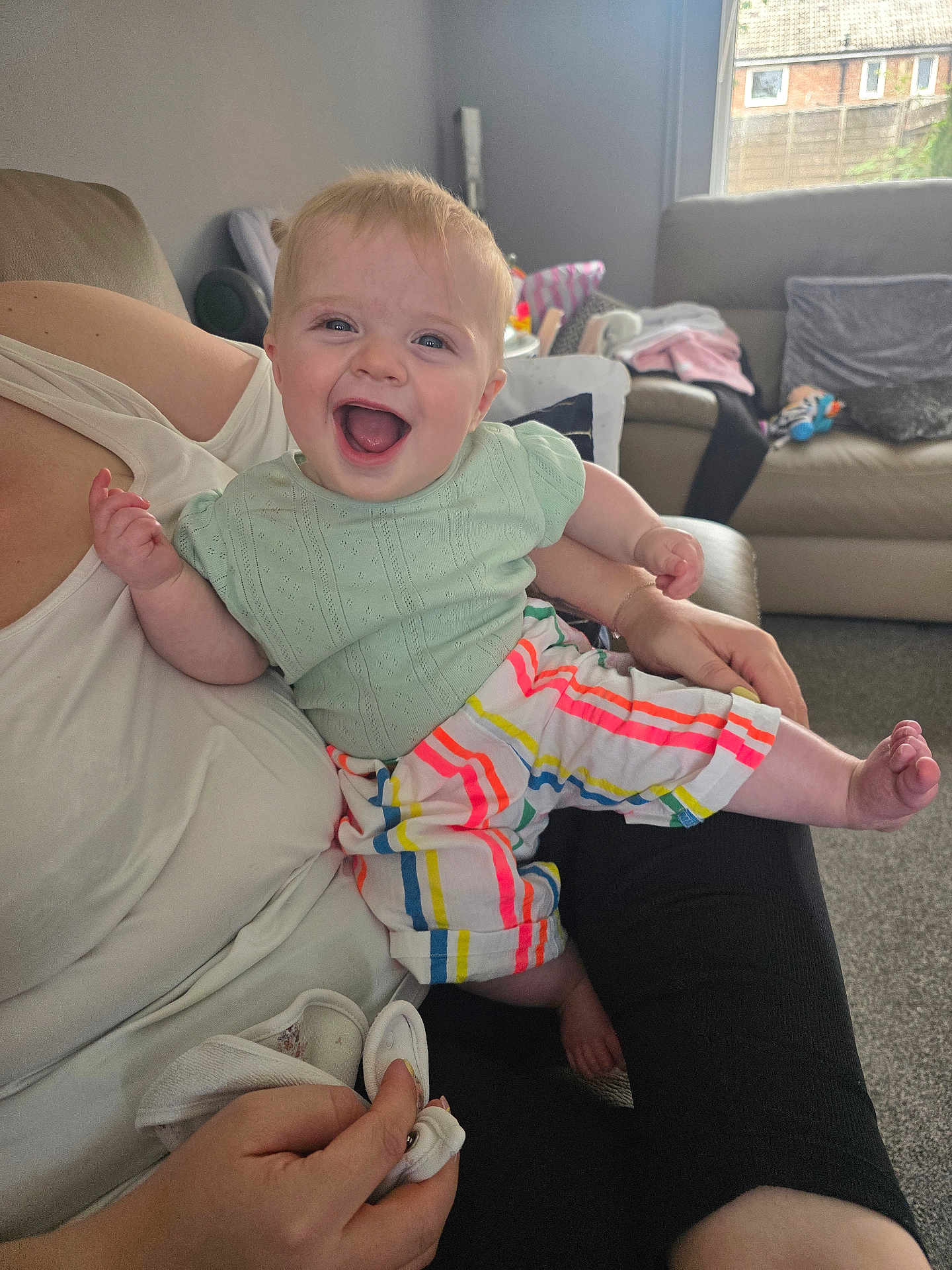 Scarlett joined the competition — help win amazing prizes! baby, child, smiling, happy, person, indoor, living_room, couch, colorful_clothing, striped_pants, mint_green_top, adult_arm, carpet, window, natural_light, furniture, casual, joy, cute, portrait