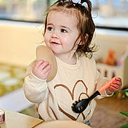 Brooklyn is registered to the contest to win money with this photo: blurry_background, child, curious, cute, happy, heart_design, home, indoor, pasta_spoon, person, pigtails, plate, playful, smile, sweater, table, toddler, toy, wooden_toy, young_child