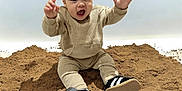 Tiago participe au concours pour gagner de l'argent avec cette photo : beige_clothing, casual_wear, child, cute, footwear, hands_up, happy, indoor, infant, light_background, person, playful, portrait, sand, sitting, smiling, sneakers, studio, toddler, young_child