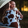 baby, woman, cowboy_hat, cow_print_jacket, outdoor, nature, portrait, holding, expression, person, forest, sunlight, warm_light, headwear, grass, skin, clothing, face, mother, child
