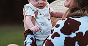 Justin joined the competition — help win amazing prizes! baby, woman, cowboy_hat, cow_print, outdoor, smiling, holding, baby_clothes, shorts, person, portrait, nature, happy, child, casual, seated, grass, daylight, family, interaction