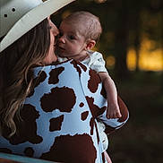 Justin is registered to the contest to win money with this photo: baby, woman, cowboy_hat, cow_print, outdoor, nature, forest, portrait, affection, holding, child, love, evening, soft_light, blurred_background, casual_clothing, head, face, person, warmth