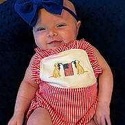 Madelyn is registered to the contest to win money with this photo: baby, child, smile, headband, blue_bow, red_stripes, onesie, embroidery, dogs, flag, cushion, indoor, portrait, cute, infant, happy, skin, hand, face, clothing