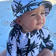 Gian Marcos is registered to the contest to win money with this photo: toddler, child, baby, hat, bucket_hat, clothing, pattern, palm_tree, outdoor, beach, blanket, sitting, portrait, cute, face, skin, expression, sunlight, summer, nature