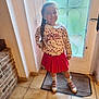 child, girl, braid, smile, indoor, door, glass, floor, tile_floor, red_skirt, floral_top, socks, shoes, mat, hand_on_hip, long_hair, pose, window, wall, cozy