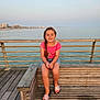 child, girl, bench, wood, sea, water, sky, braid, pink_top, denim_shorts, sandals, smiling, outdoor, daylight, coast, pier, railing, relaxation, vacation, nature