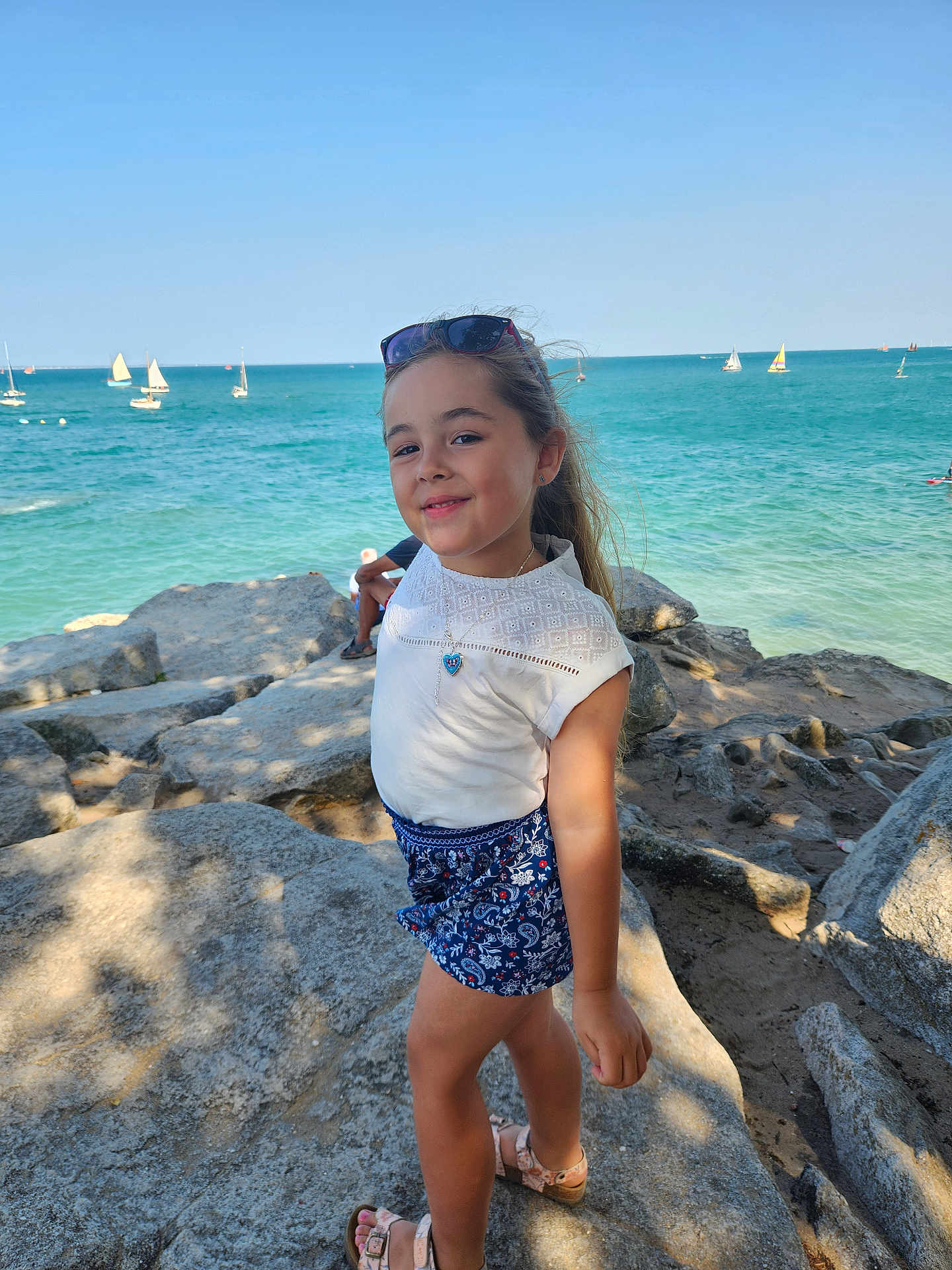 Mia a rejoint le concours — aidez-le/la à gagner de superbes lots ! child, girl, smiling, rock, ocean, sea, sailboat, blue_sky, sunlight, summer, shore, casual_clothing, sandals, necklace, sunglasses, outdoor, nature, vacation, happy, portrait