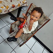 Kyrin is registered to the contest to win money with this photo: toddler, child, portrait, smiling, sitting, snacking, food, snack_packet, chair, indoor, kitchen, table, tablecloth, autumn_leaves, sneakers, clothing, curly_hair, tile_floor, wall, happy
