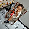 toddler, child, portrait, smiling, sitting, snacking, food, snack_packet, chair, indoor, kitchen, table, tablecloth, autumn_leaves, sneakers, clothing, curly_hair, tile_floor, wall, happy