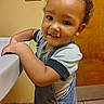 Kyrin joined the competition — help win amazing prizes! child, toddler, baby, smiling, portrait, indoor, bathroom, sink, overalls, shirt, curly_hair, face, eyes, teeth, standing, leaning, wooden_door, wall, tile_floor, light_switch