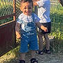 child, boy, smiling, outdoor, sunlight, shadow, denim_overalls, tshirt, sandals, grass, metal_gate, fence, nature, play, summer, happy, two_children, portrait, daylight, casual_clothing