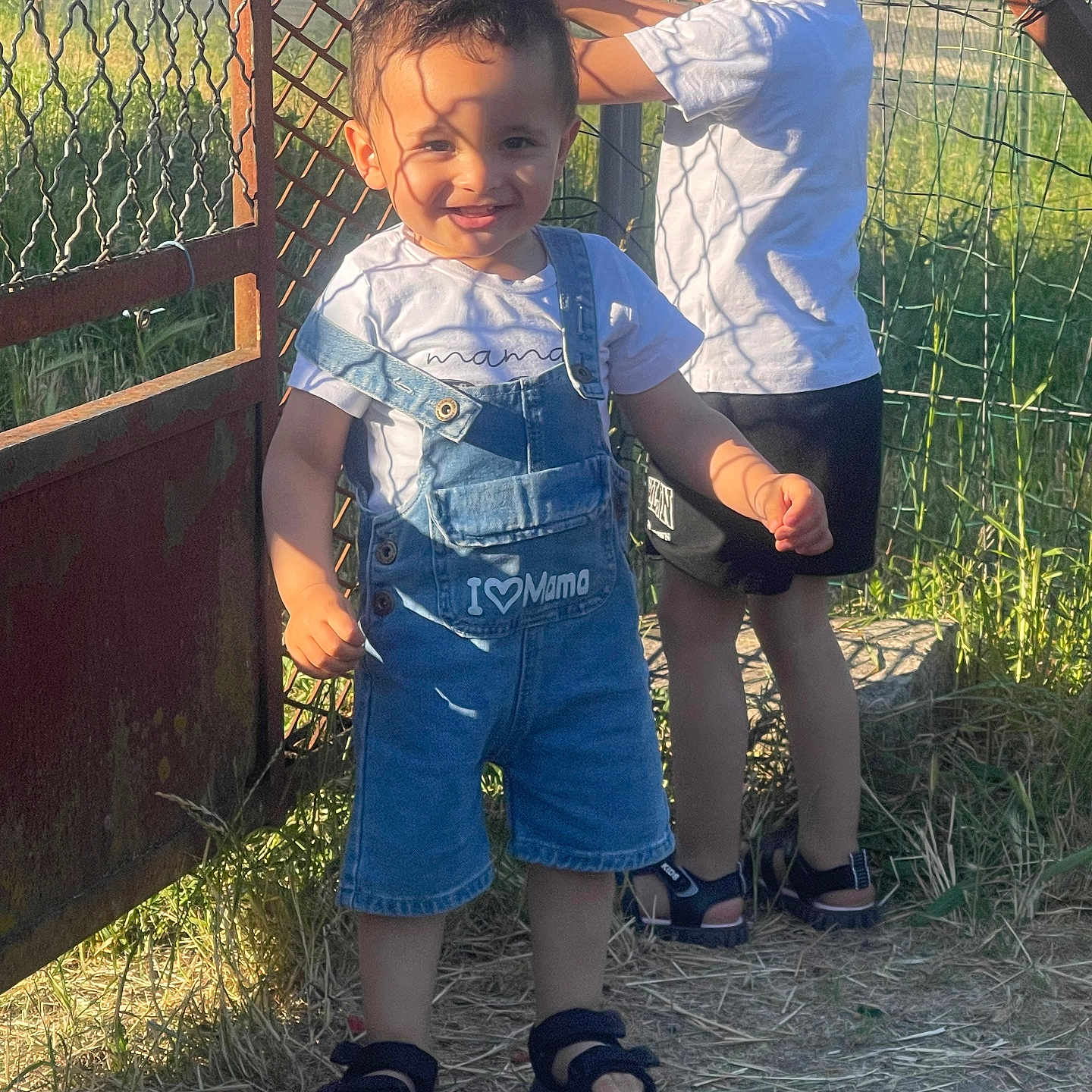 Iyaas a rejoint le concours — aidez-le/la à gagner de superbes lots ! boy, casual_clothing, child, daylight, denim_overalls, fence, grass, happy, metal_gate, nature, outdoor, play, portrait, sandals, shadow, smiling, summer, sunlight, tshirt, two_children